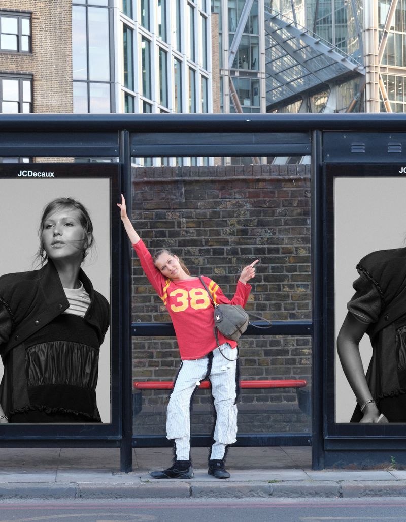 Angelina Kendall posing playfully inside a bus shelter between two large black-and-white fashion billboards on a city street.