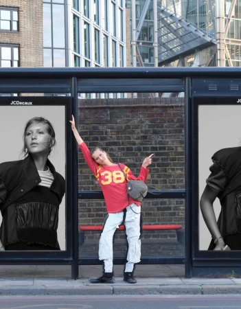 Angelina Kendall posing playfully inside a bus shelter between two large black-and-white fashion billboards on a city street.