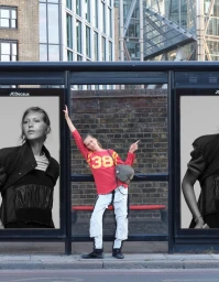 Angelina Kendall posing playfully inside a bus shelter between two large black-and-white fashion billboards on a city street.