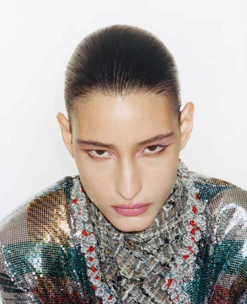 Close-up beauty portrait of model Luiza Perote with slicked-back hair, graphic pink eyeliner, and metallic textured jacket on white.