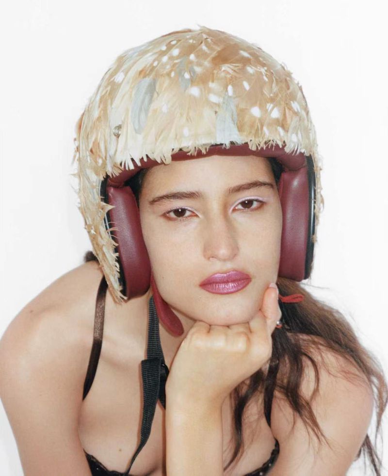 Model Luiza Perote wearing a feather-covered helmet with burgundy interior padding resting her chin on her hand against a white background.