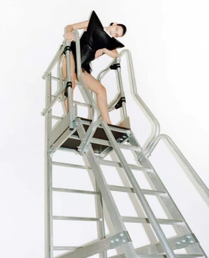 Model Luiza Perote in a sculptural black top posing at the top of a tall silver metal ladder against a white background.