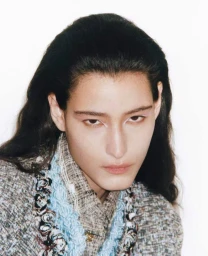 Close-up portrait of a model with slicked-back dark hair wearing a tweed garment against a white background.