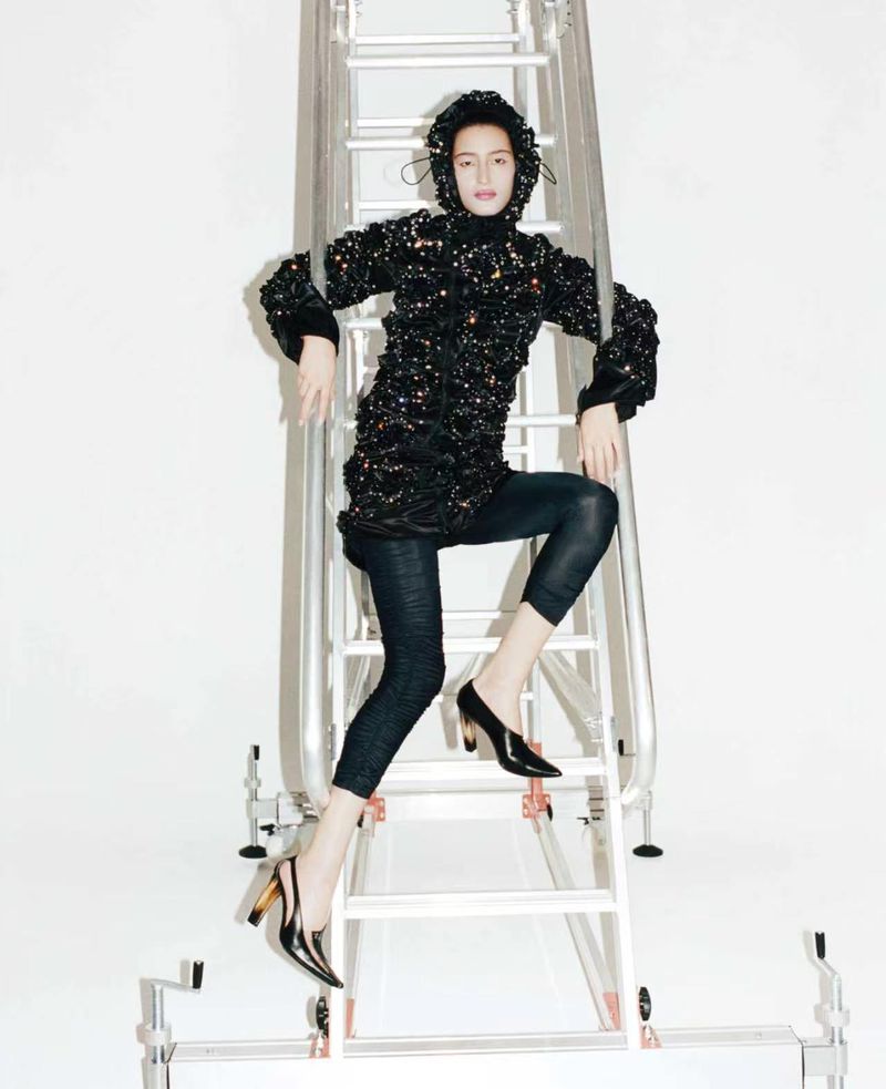 Model Luiza Perote poses on an aluminum studio ladder wearing a black textured hooded jacket, cropped tights, and black pointed heels.