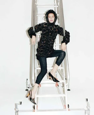 Model Luiza Perote poses on an aluminum studio ladder wearing a black textured hooded jacket, cropped tights, and black pointed heels.
