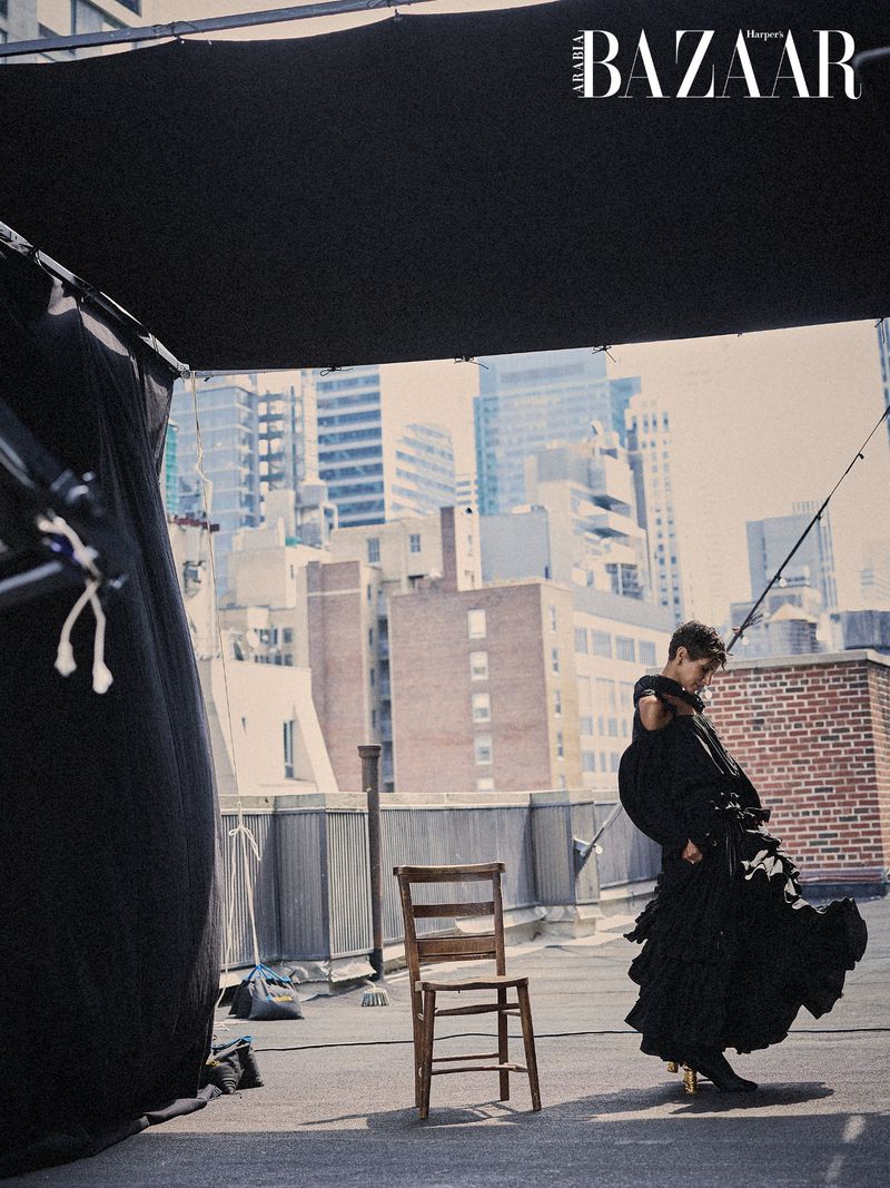 Deena Aljuhani Abdulaziz in a voluminous black ruffled dress poses on an urban rooftop with a wooden chair.