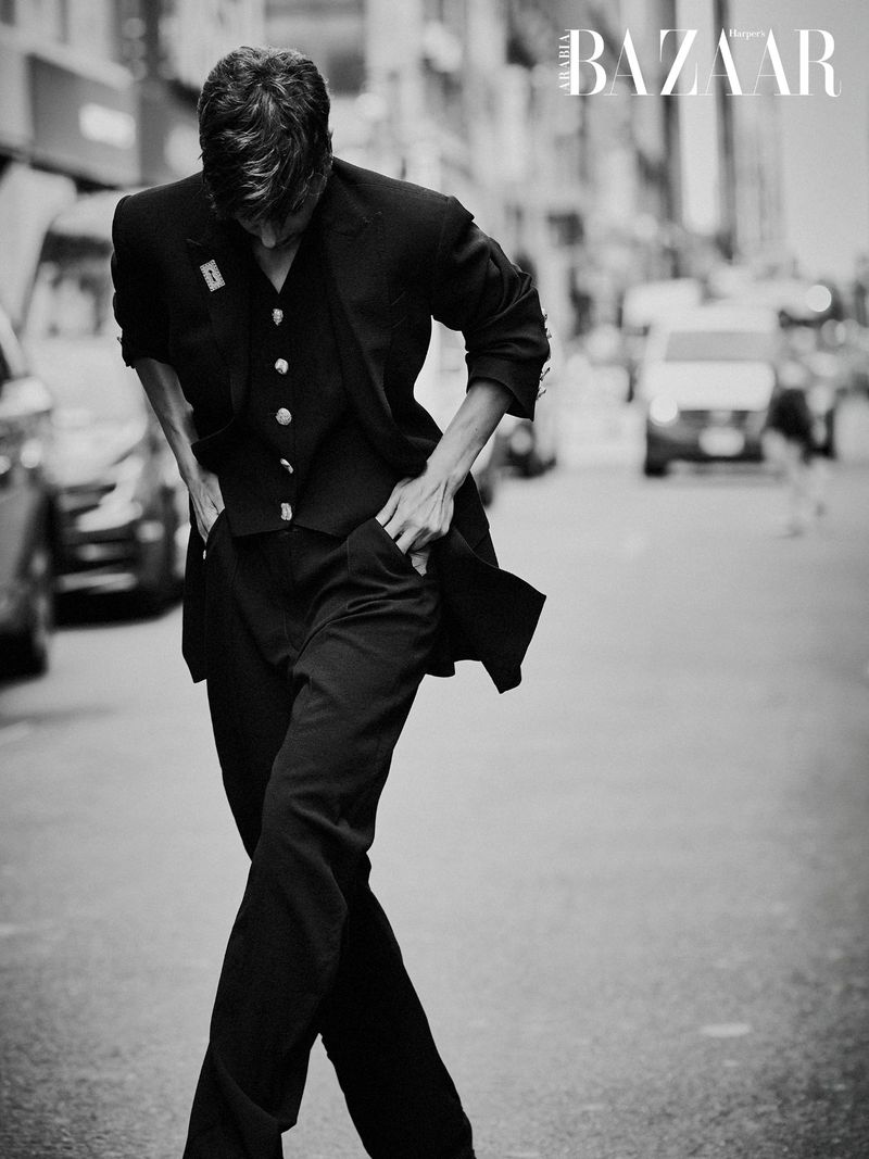 Black and white editorial photograph of Deena Aljuhani Abdulaziz walking down a city street in a tailored black three-piece suit.