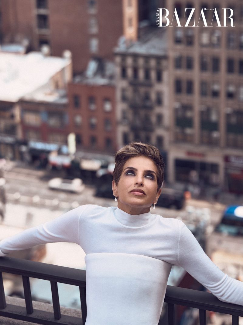 Deena Aljuhani Abdulaziz looking upward while leaning on a balcony railing in a white long-sleeve top against a city backdrop.