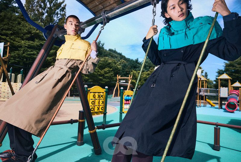 Two male models in color-blocked trench coats posing on playground swings against a bright sunny outdoor backdrop.