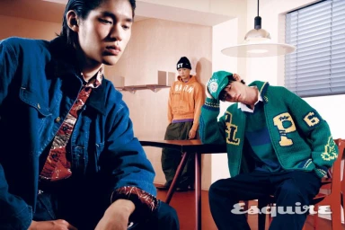 Three young male models posing in menswear including a blue denim jacket, green varsity cardigan, and orange hoodie in an interior set.