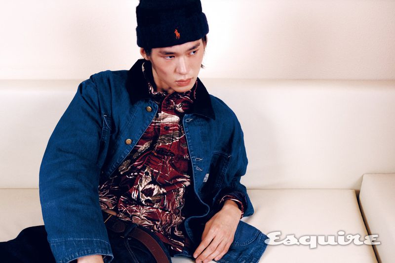 Male model wearing a blue denim jacket, red patterned shirt, and dark knit beanie seated on an off-white leather couch.