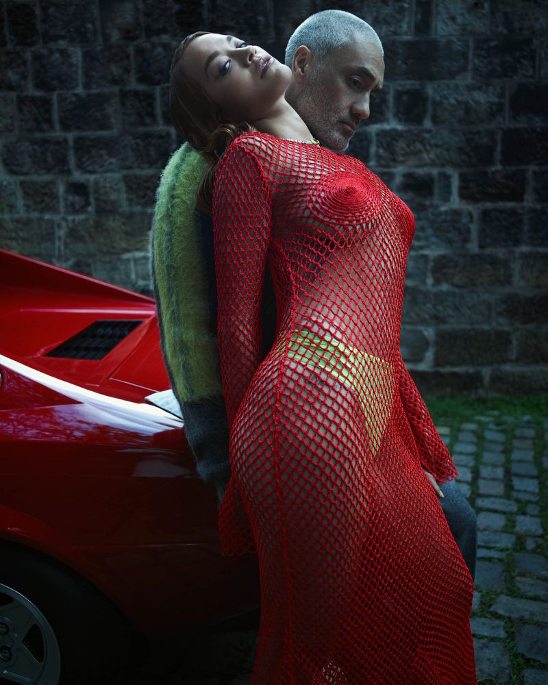 A woman in a red mesh dress leans against a red car, with a man behind her against a brick wall; outdoor setting with intimate pose.