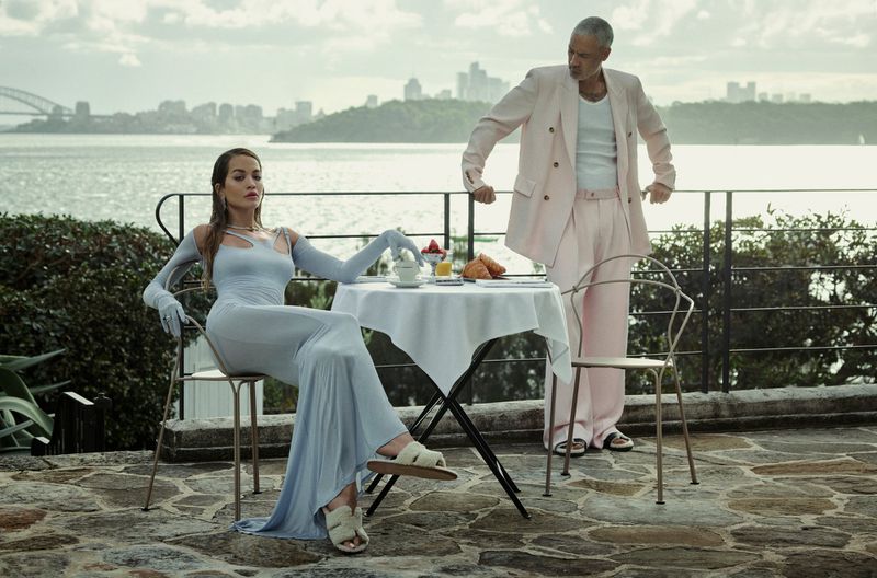 Rita Ora seated at a table outdoors wearing a light blue dress with matching gloves while a man stands nearby in a pink suit