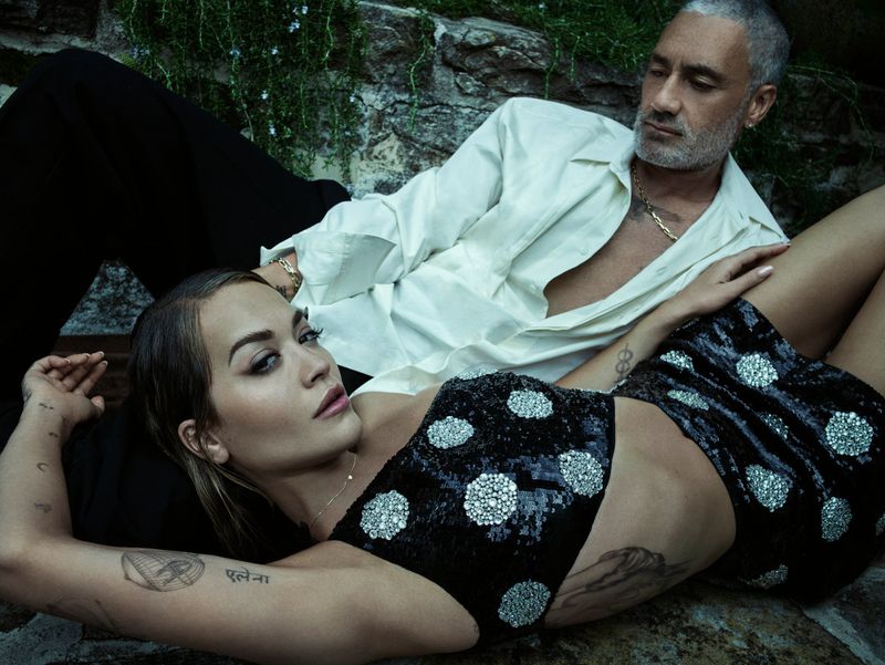 Rita Ora and Taika Waititi are laying outdoors on stone, photographed in a close up studio shot. Ora is wearing a black sequin crop top.