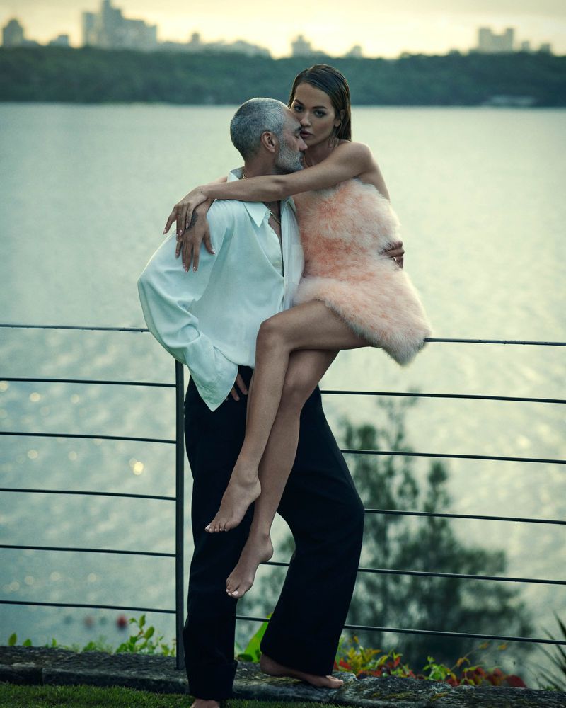 An adult male carrying an adult female near a body of water, with a city skyline visible in the background.