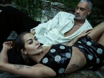Rita Ora in a black sequined top resting her head on a man in a white shirt outdoors against a stone wall