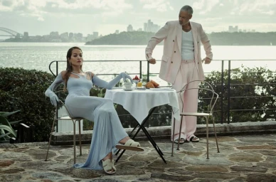 Rita Ora seated at a table outdoors wearing a light blue dress with matching gloves while a man stands nearby in a pink suit