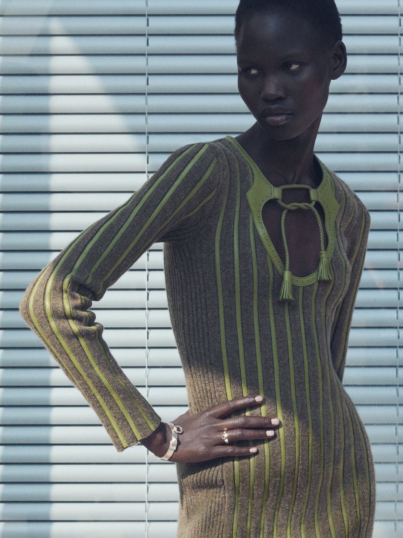 Woman in brown ribbed midi dress with green accents posing in front of blinds in studio. Fashion editorial, portrait, neutral colors.