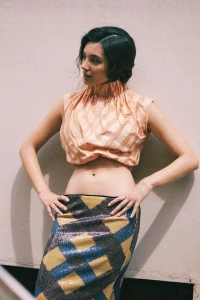 Mariasole Pollio posing with hands on hips wearing a peach high-neck crop top and sequined plaid midi skirt against a pale wall.
