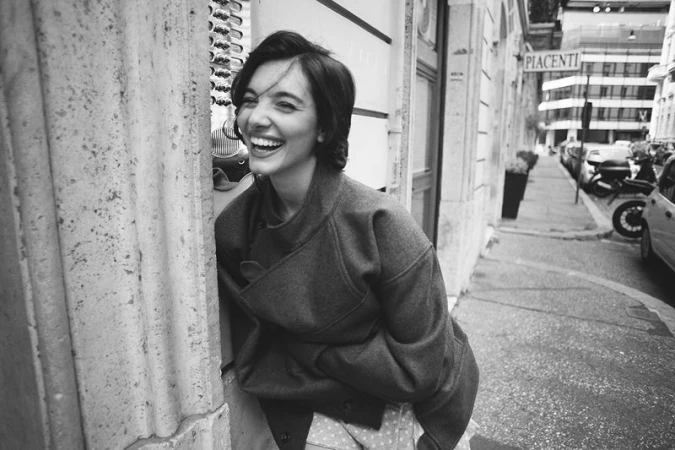 Black and white photo of a smiling woman in an oversized coat leaning against a stone doorway on a city sidewalk.