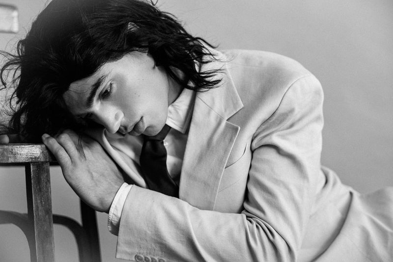 Black and white portrait of a young man with long dark wavy hair resting his head on a wooden stool, wearing a light suit, white shirt, and dark tie.