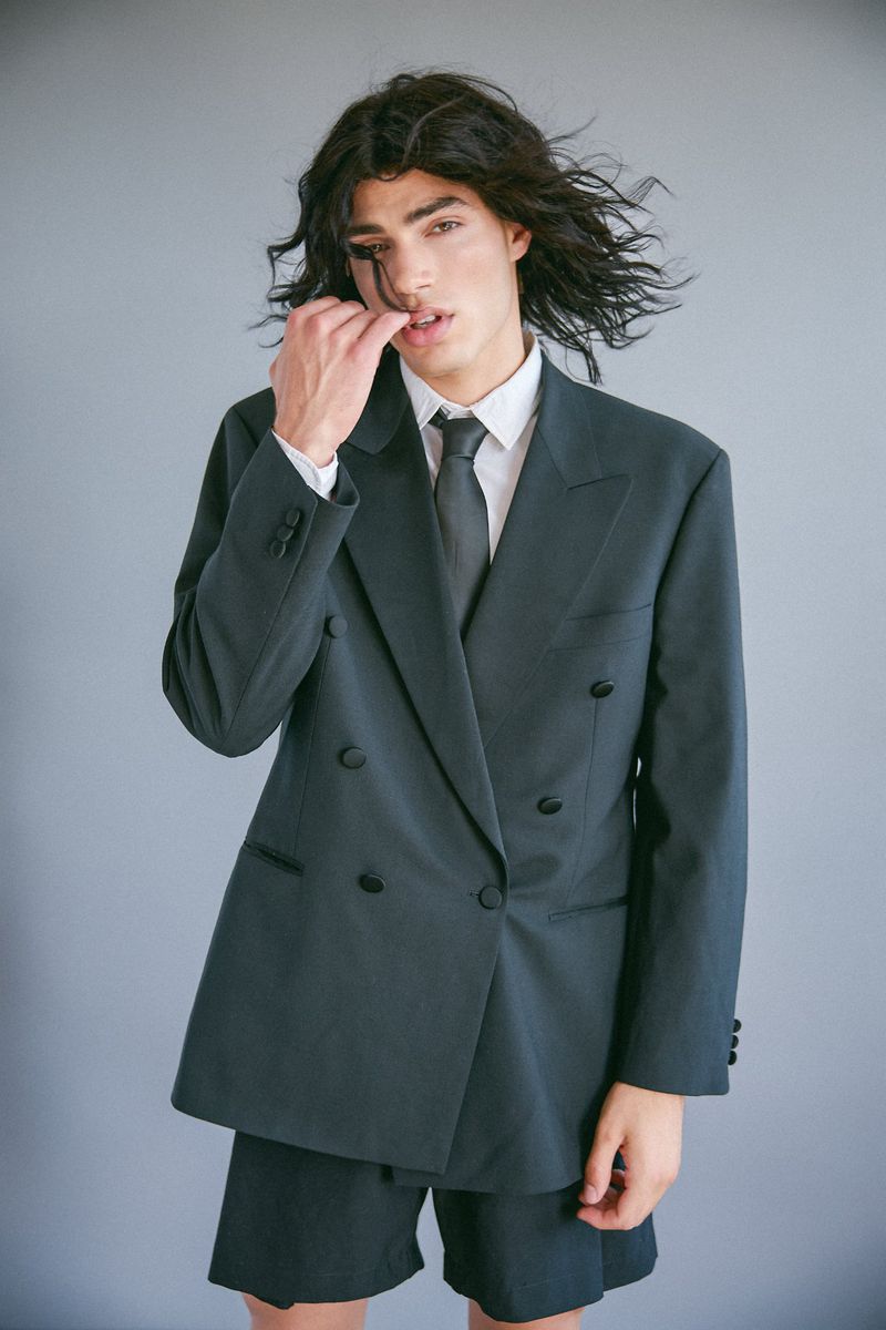 Model Ruben Aires wearing an oversized dark double-breasted suit jacket with shorts and a tie against a grey backdrop.