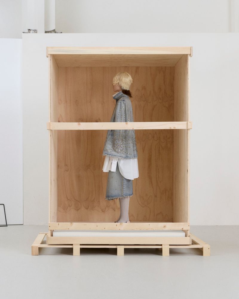 Model in a denim outfit standing in profile inside a large open wooden shipping crate against a white studio background.