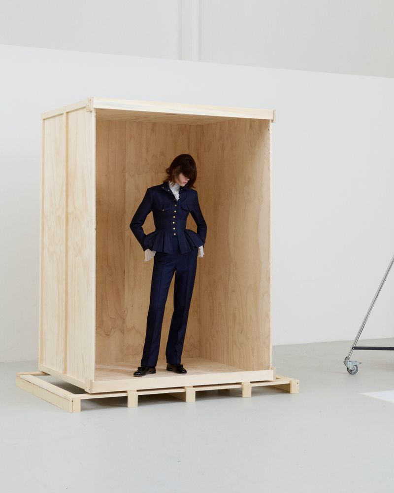 Model in a navy blue peplum suit standing inside a large open wooden shipping crate on a pallet against a white studio background.