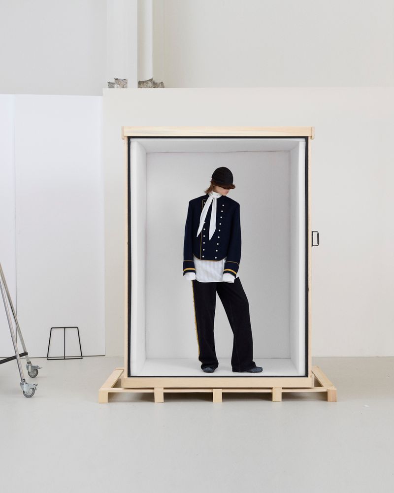 Model wearing a navy military jacket and black trousers stands inside a large wooden shipping crate in a white studio.