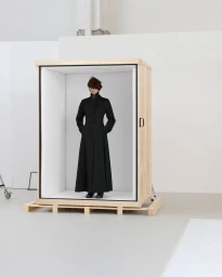 Model wearing a long black coat standing inside an open wooden shipping crate placed on a pallet in a white studio.