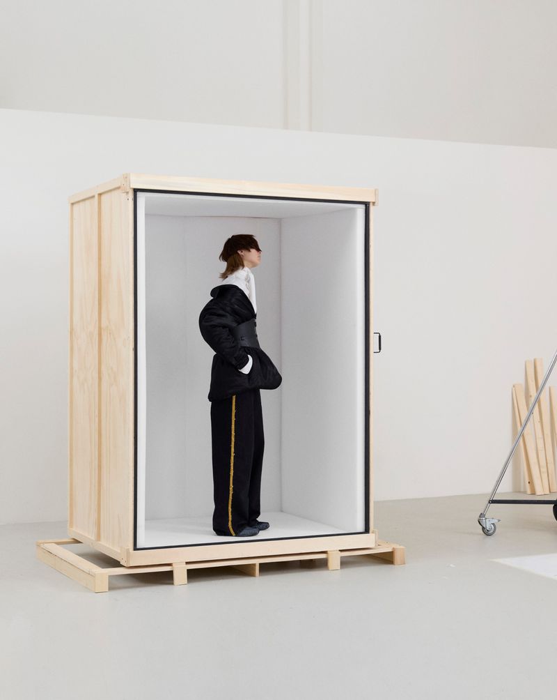 A model wearing a black cinched jacket and side-striped trousers standing inside an open wooden shipping crate in a studio.