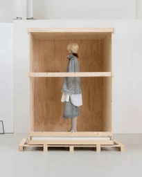 Model in a grey denim ensemble standing inside a large open wooden shipping crate inside a minimalist white studio.