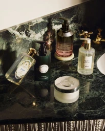 Luxury skincare and fragrance bottles arranged on a dark green marble bathroom vanity with gold fixtures.