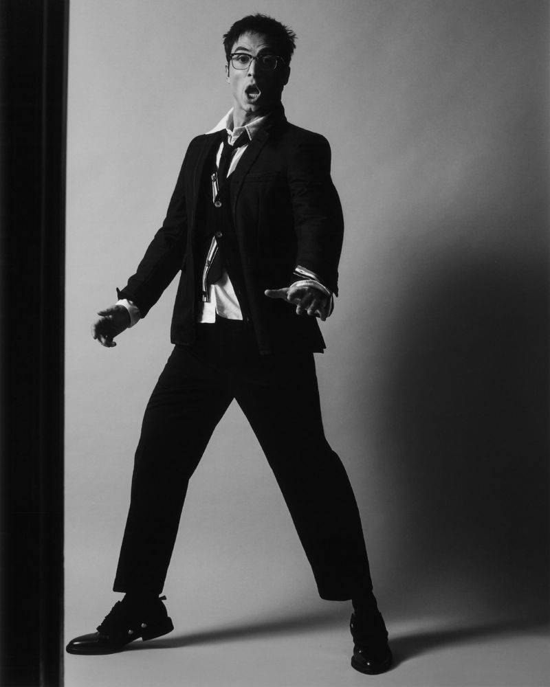 Benito Skinner posing expressively in a dark tailored suit, unbuttoned collared shirt, and glasses against a studio backdrop.