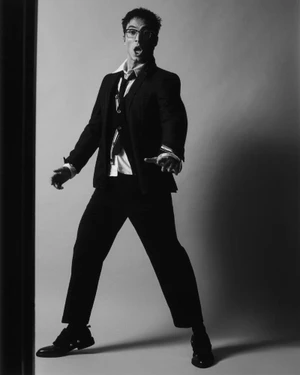 Benito Skinner posing expressively in a dark tailored suit, unbuttoned collared shirt, and glasses against a studio backdrop.