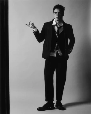 Benito Skinner posing in a black suit with a loosened tie and eyeglasses against a studio backdrop.