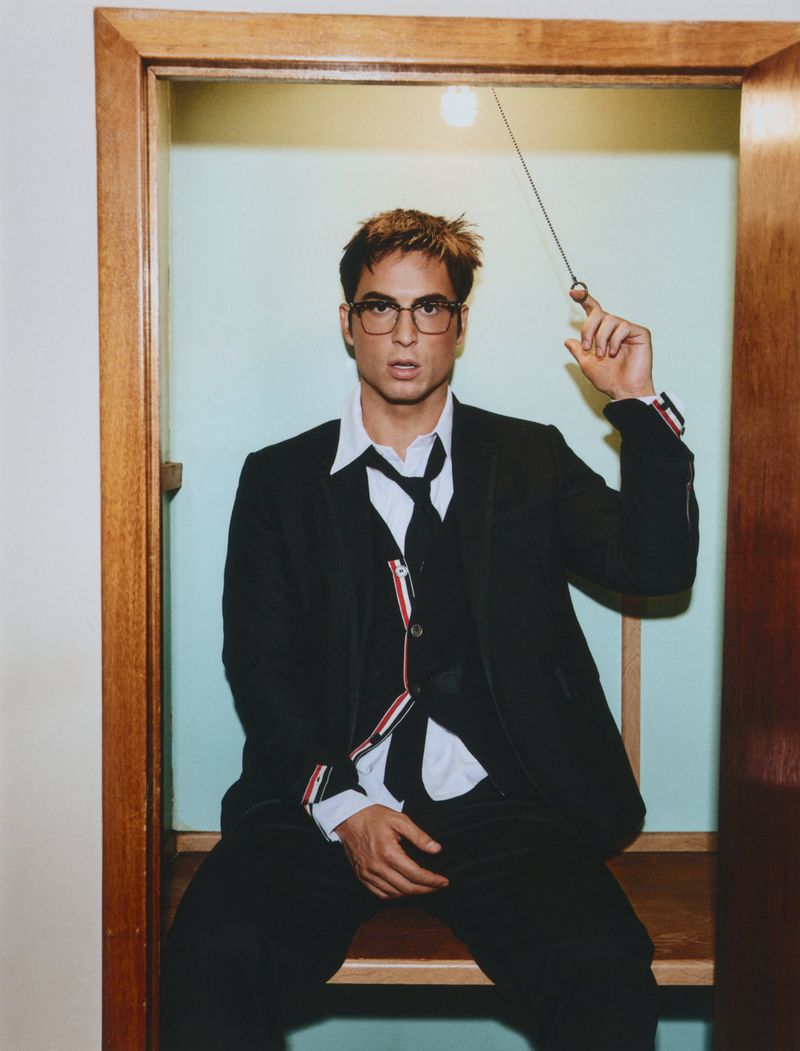 Benito Skinner wearing eyeglasses and a black suit with a loosened tie, seated inside a wooden frame holding an overhead light string.