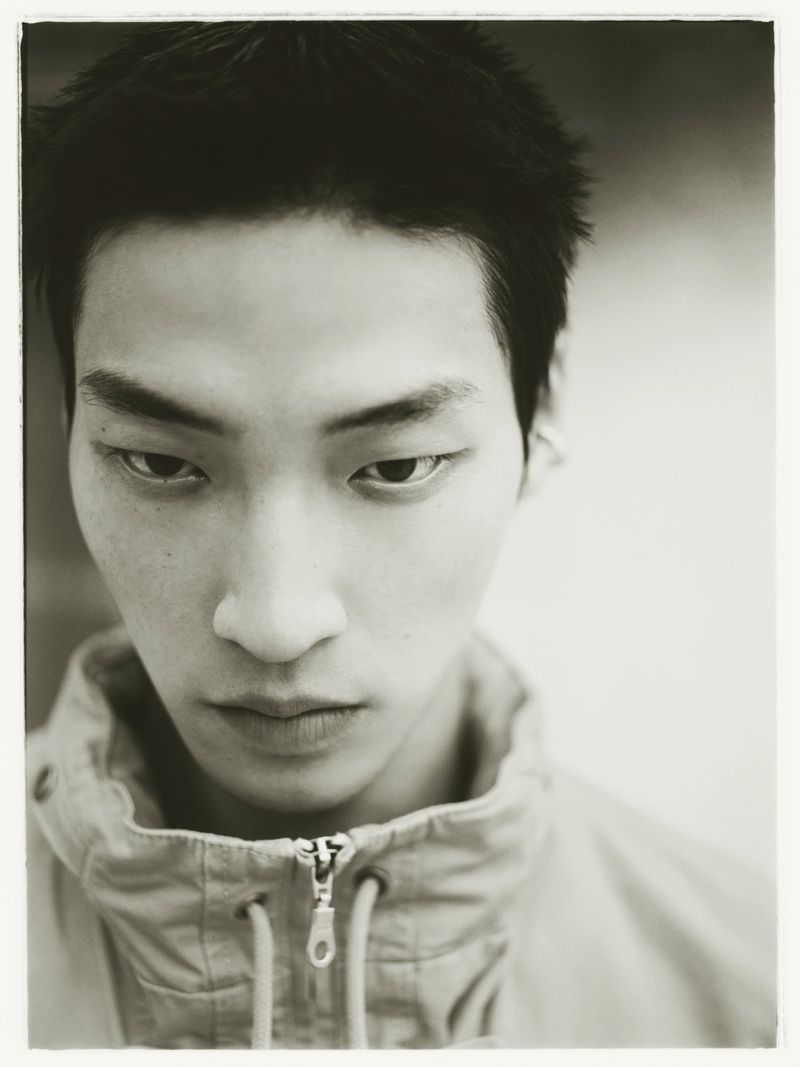Close-up monochrome portrait of an East Asian young man wearing a high-collar zip-up jacket looking down at the camera.
