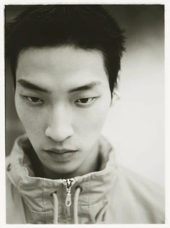 Close-up monochrome portrait of an East Asian young man wearing a high-collar zip-up jacket looking down at the camera.