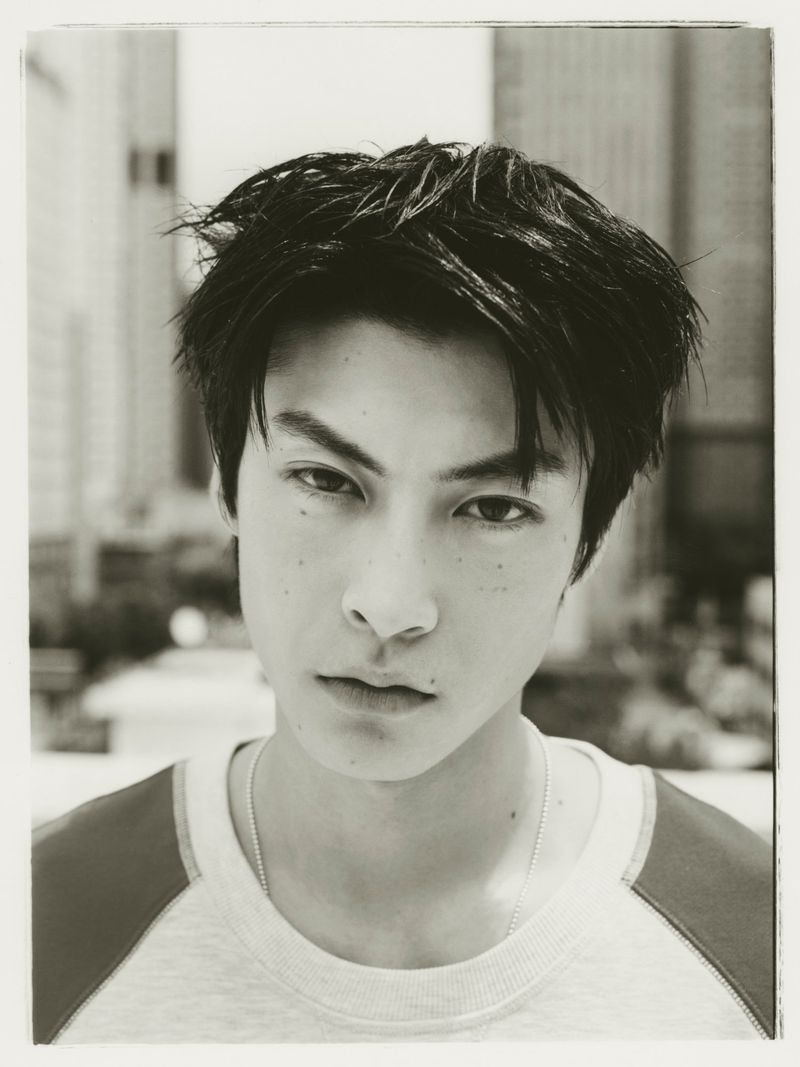 Black and white close-up portrait of an East Asian man wearing a crewneck raglan shirt with blurred city buildings in the background.