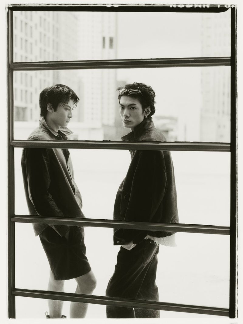 Two male models in dark casual wear pose behind a horizontal metal grid structure with urban buildings in the background.