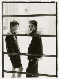 Two male models in dark casual wear pose behind a horizontal metal grid structure with urban buildings in the background.