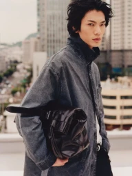 East Asian male model holding a black leather bag and wearing a faded grey jacket on a city rooftop.