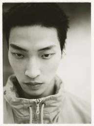 Close-up monochrome portrait of an East Asian young man wearing a high-collar zip-up jacket looking down at the camera.