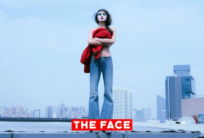Model posing on an urban rooftop wearing flared blue denim jeans and holding a red jacket across her torso against a cityscape skyline.