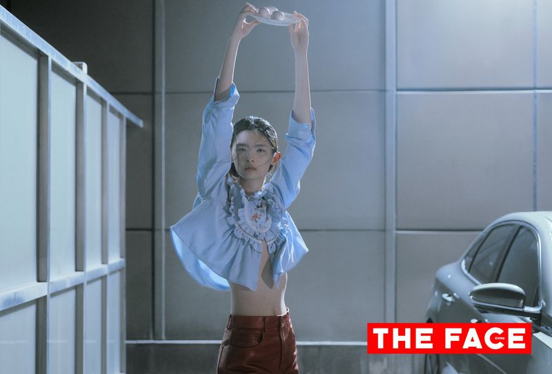 Model in a cropped blue ruffled shirt and brown leather pants holding a plate with eggs overhead next to a car.