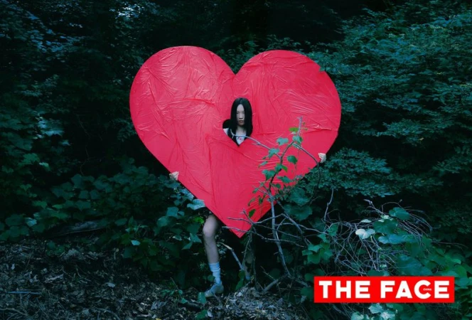Model looking through an opening in a giant red textured heart prop amidst dense green forest foliage.