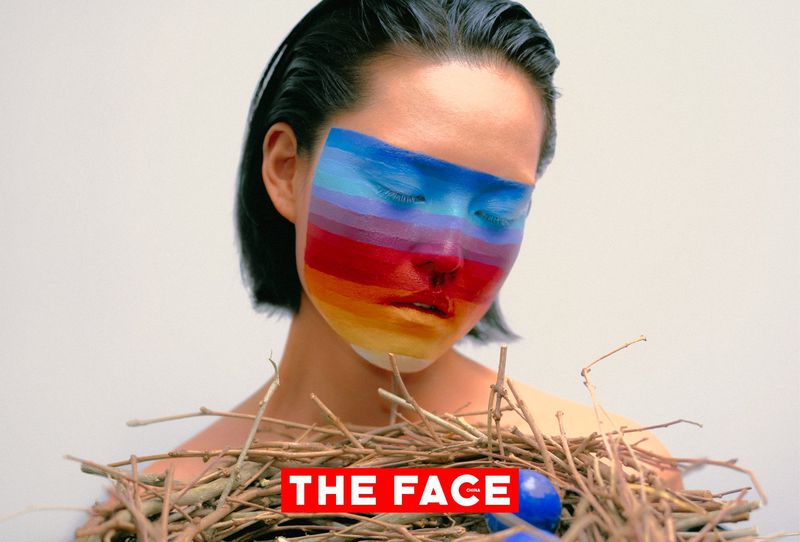 East Asian model with rainbow face paint looking down over a twig nest with blue eggs against a white background.
