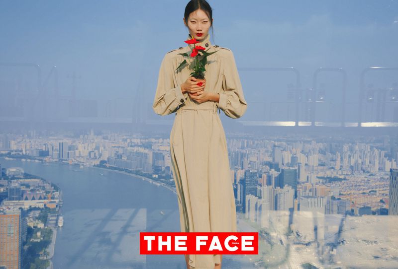 Model in a beige belted trench dress holding a potted red flower in front of a high-rise window overlooking a city skyline.