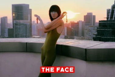 Model in an olive jumpsuit holding red beaded strings against a city skyline at sunset.